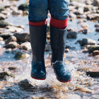 A person wearing 'Classic Wellies Navy' is jumping in a stream, creating a splash. Rocks surround the water and red socks show above the wellies.